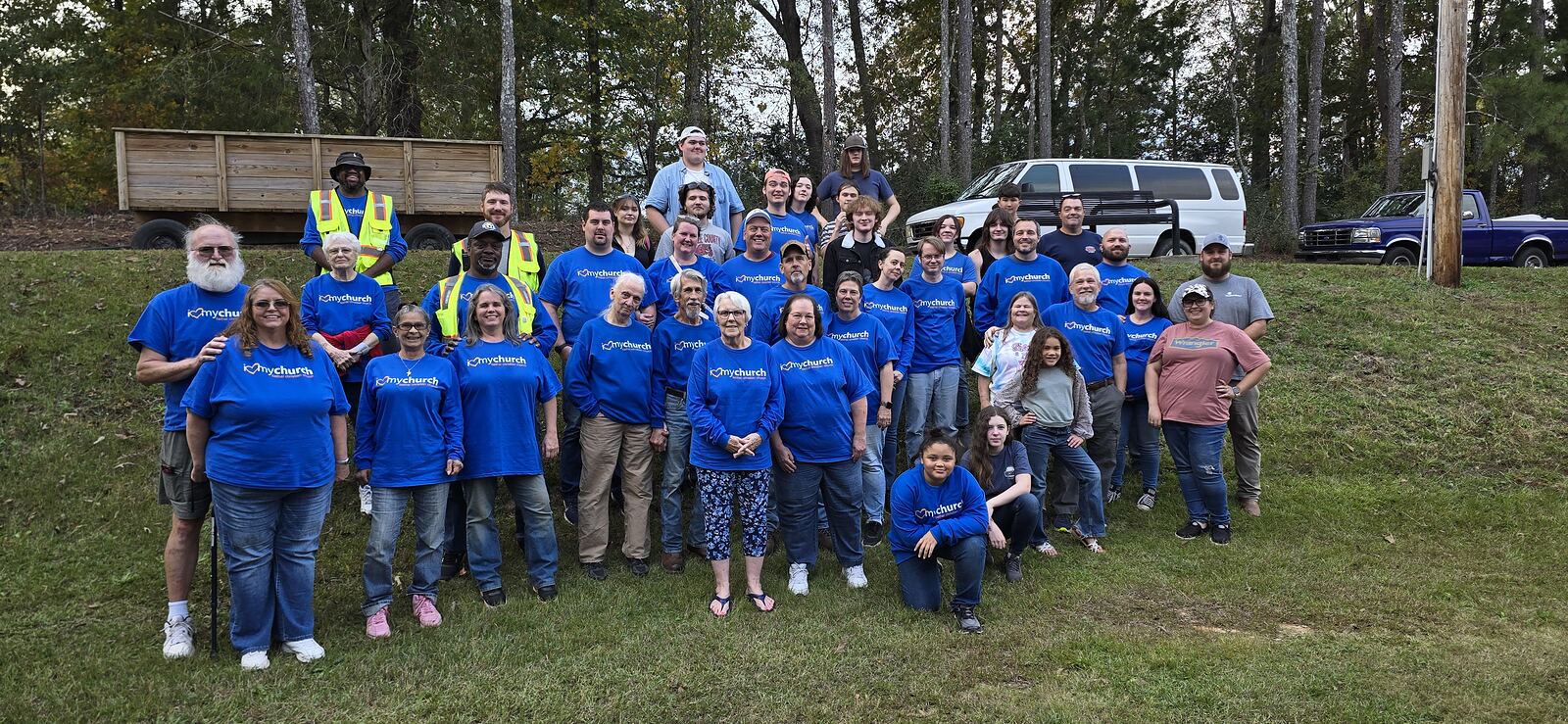 Bethel volunteers gathered on the grass at the 2024 Fall Festival, most wearing royal blue shirts with the mychurch logo across the chest &mdash; both short-sleeved and long-sleeved styles are visible